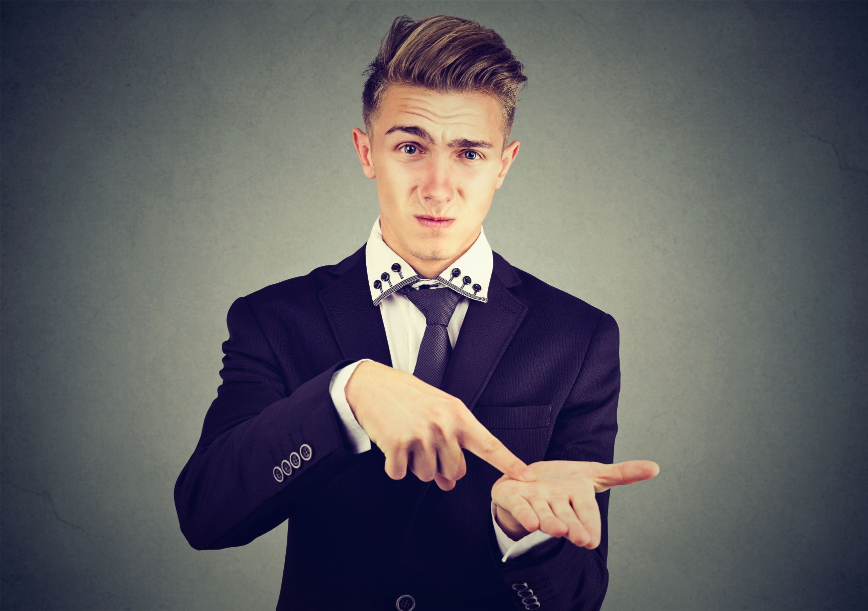 Young man in suit stares into camera and points to his empty hand, demanding money.